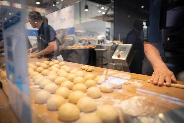 Reihen aus Teigkugeln auf bemehltem Holztisch hinter Glas; Hand rollt Teig, Bäcker im Hintergrund.