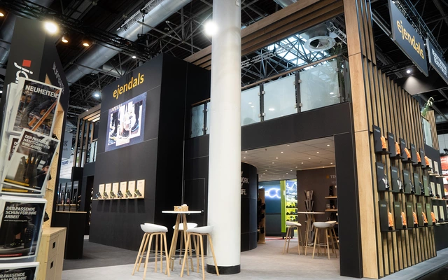 Messestand mit schwarzem und holzfarbenem Design, hohen Stühlen und Tischen, beleuchtet durch Deckenlampen in einer Messehalle.