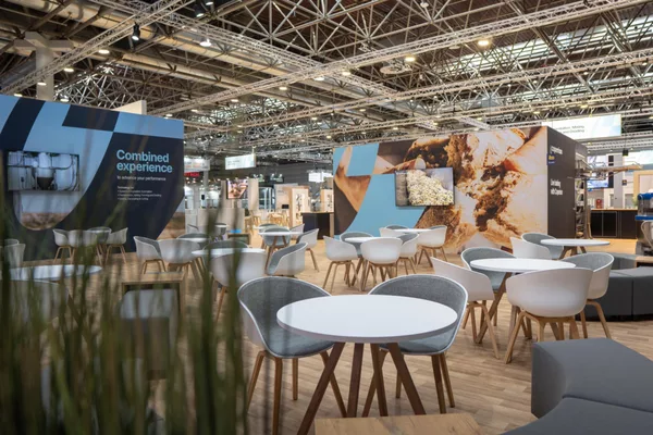 Loungebereich mit vielen runden weißen Tischen und Stühlen, Holzfußboden, Wandgrafiken „Combined experience“ und Teigmotiv.