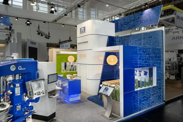 [Translate to English:] Detailansicht des Niemann-Messestands mit Lounge-Bereich, blauen Wandgrafiken und interaktivem Terminal.