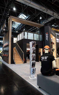 Messestand mit zweigeschossigem Aufbau, Treppe, Prospektständer und Schaufensterpuppe mit Schutzhelm und T-Shirt mit Aufdruck im Bereich Arbeitssicherheit.