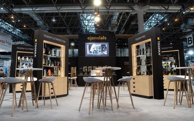 Messestand von ejendals mit Holztheken und hohen Stühlen, umgeben von Regalen mit Schutzhandschuhen, in einer Messehalle mit sichtbarer Deckenbeleuchtung.