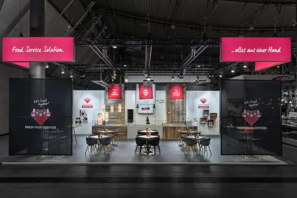 Übersicht über den offenen Messestand mit Sitzgruppen, Vitrinen und leuchtenden Deckentransparenzen.