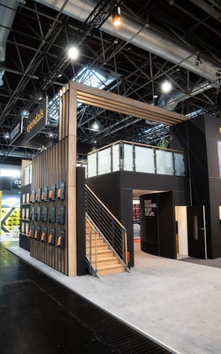 Messestand mit zweistöckiger Konstruktion aus Holz und schwarzen Elementen, Treppe mit Holzstufen, mehrere orangefarbene Sicherheitshandschuhe an der Außenwand befestigt, Deckenbeleuchtung und sichtbare Rohrleitungen in der Halle.
