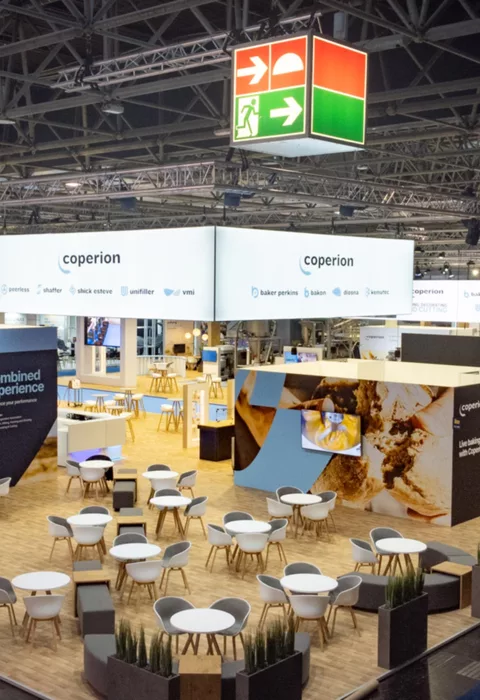 Top view of exhibition stand with many round tables, large ceiling banners “coperion” and baking area with oven station