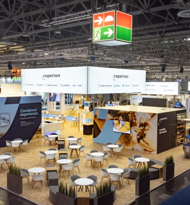 Top view of exhibition stand with many round tables, large ceiling banners “coperion” and baking area with oven station