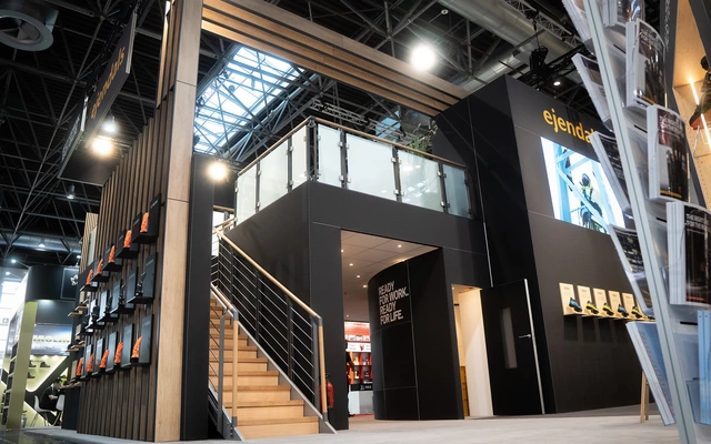 Messestand mit zweistöckigem Aufbau, Holztreppe, Glasgeländer und schwarzer Verkleidung auf einer Messe für Arbeitssicherheit.