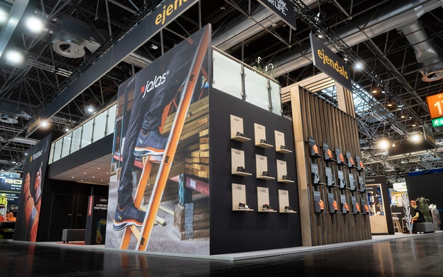 Messestand mit großem Bild einer Person auf einer Leiter, Wand mit Holzpaneelen und mehreren kleinen Schildern, Deckenbanner mit der Aufschrift 'ejendals' in einer Messehalle mit sichtbarer Beleuchtung und anderen Ständen im Hintergrund.