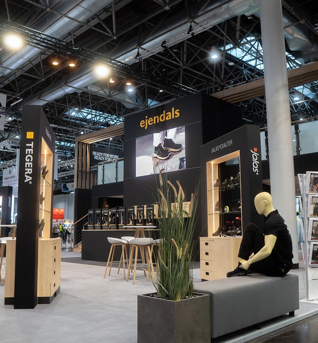 Messestand mit der Marke ejendals auf einer Messe für Arbeitssicherheit, mit Sitzbank, Schaufensterpuppe in schwarzer Kleidung und Produktpräsentationen an den Wänden.