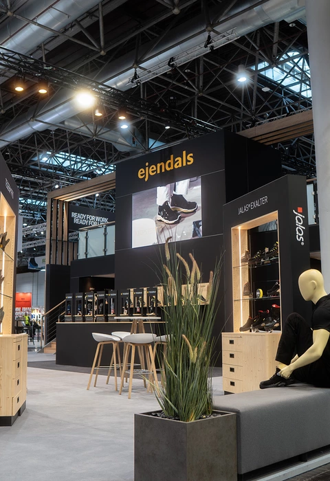 Messestand mit der Marke ejendals auf einer Messe für Arbeitssicherheit, mit Sitzbank, Schaufensterpuppe in schwarzer Kleidung und Produktpräsentationen an den Wänden.