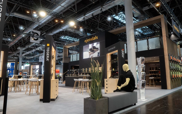 Messestand mit der Marke ejendals auf einer Messe für Arbeitssicherheit, mit Sitzbank, Schaufensterpuppe in schwarzer Kleidung und Produktpräsentationen an den Wänden.