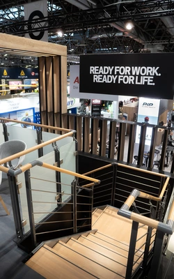 Treppenaufgang mit Holzgeländer und Metallstreben auf einer Messe mit einem großen Schild mit der Aufschrift 'READY FOR WORK. READY FOR LIFE.'